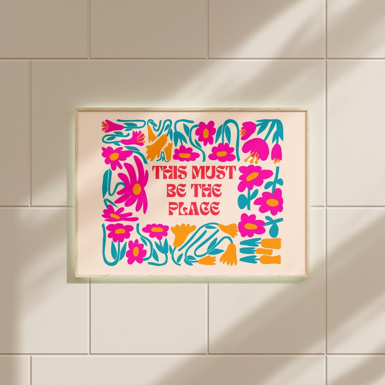 以下が含まれることがあります： ピンクとターコイズブルーの花柄プリントに、赤い文字で「This must be the place」と書かれています。プリントは明るい木製のフレームで額装されています。