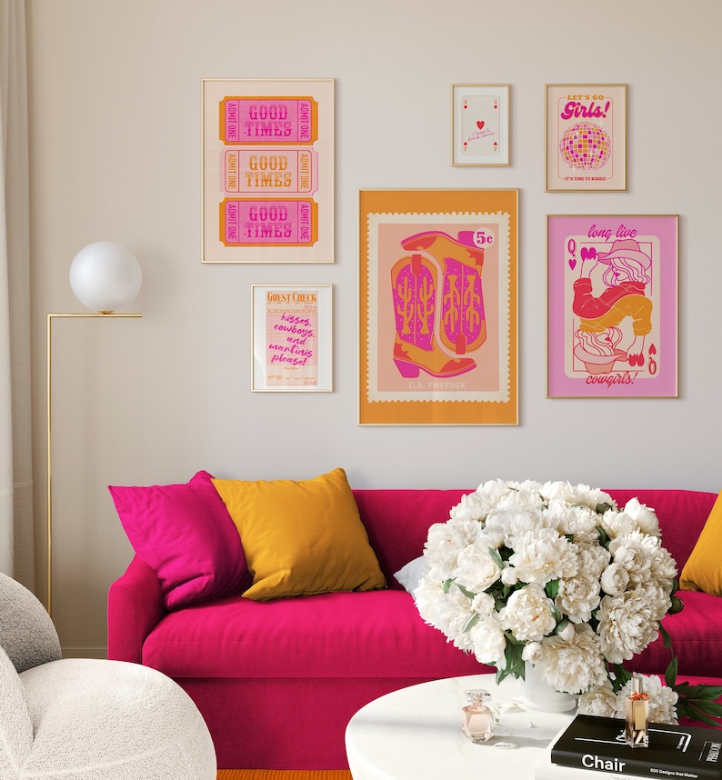 May include: A pink sofa with yellow and pink pillows in a living room with a white flower arrangement on a white coffee table. The wall behind the sofa is decorated with a collection of framed prints with a western theme, including a cowboy boot, a queen of hearts playing card, and a sign that says "Good Times".
