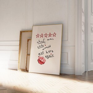 Op de afbeelding: Een witte poster met een gouden lijst met een vijfsterrenbeoordeling en de tekst "kok was schattig! zou hier weer eten" met een rode lippenstiftkus.