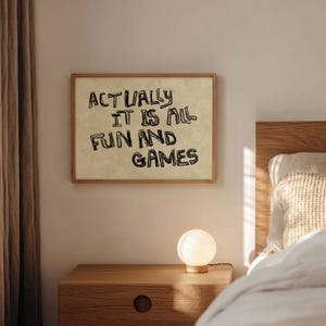 Pode incluir: Uma impress&atilde;o emoldurada com o texto "ACTUALLY IT IS ALL FUN AND GAMES" em letras pretas. A impress&atilde;o est&aacute; pendurada acima de uma mesa de cabeceira de madeira com uma pequena l&acirc;mpada redonda e iluminada. A cena &eacute; ambientada em um quarto com uma paleta de cores neutras.