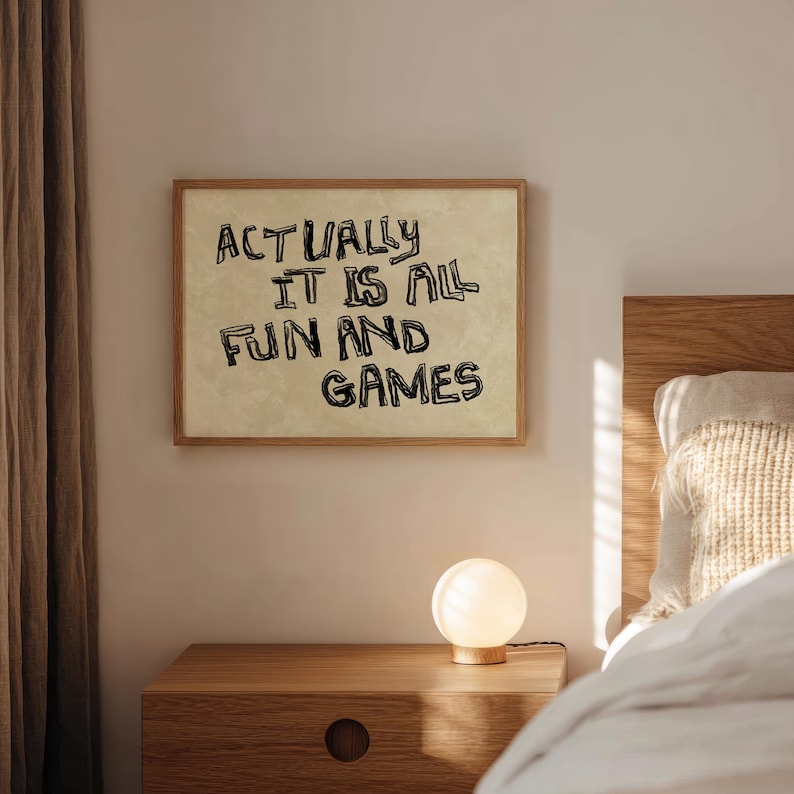 Pode incluir: Uma impress&atilde;o emoldurada com o texto "ACTUALLY IT IS ALL FUN AND GAMES" em letras pretas. A impress&atilde;o est&aacute; pendurada acima de uma mesa de cabeceira de madeira com uma pequena l&acirc;mpada redonda e iluminada. A cena &eacute; ambientada em um quarto com uma paleta de cores neutras.