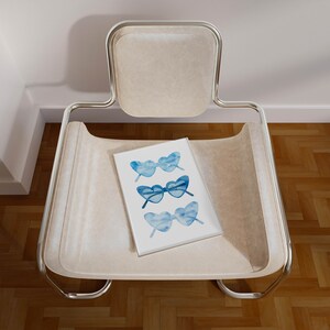 May include: A white chair with a chrome frame and a white cushion. A white card with a blue watercolor illustration of three pairs of sunglasses is on the cushion.