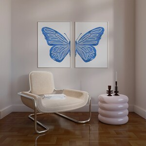 May include: A light beige armchair with a chrome frame sits in a room with a light pink side table and two candlesticks. Two framed prints of a blue butterfly are on the wall behind the chair.