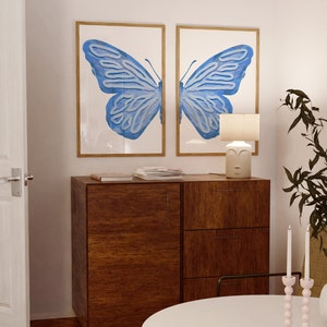 May include: Two framed prints of a blue butterfly with a wooden cabinet and a white table lamp with a face on the base. The table lamp is on a wooden cabinet with two drawers. There are two pink candle holders with white candles on the table.