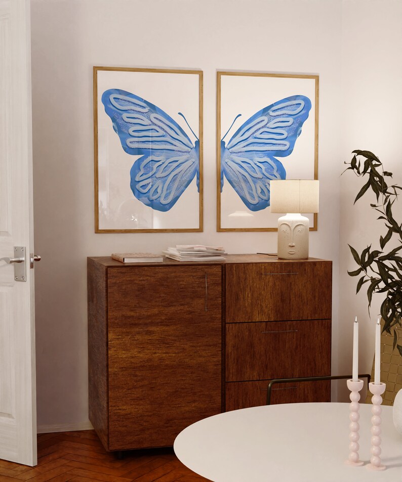 May include: Two framed prints of a blue butterfly with a wooden cabinet and a white table lamp with a face on the base. The table lamp is on a wooden cabinet with two drawers. There are two pink candle holders with white candles on the table.