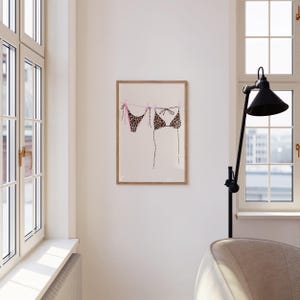 May include: A framed print of a leopard print bikini hanging on a clothesline with pink clothespins. The print is on a white background.