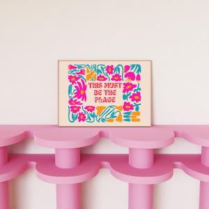 以下が含まれることがあります： クリーム色のプリントで、ピンク、ブルー、イエローの色の花柄の縁取りがされています。テキスト「THIS MUST BE THE PLACE」はピンクとブルーです。