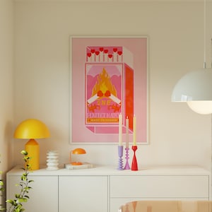 May include: A pink and orange poster with a matchbox illustration. The matchbox has a red flame and the text "ONE PERFECT MATCH MADE IN HEAVEN". Three candlesticks with candles are in front of the poster.