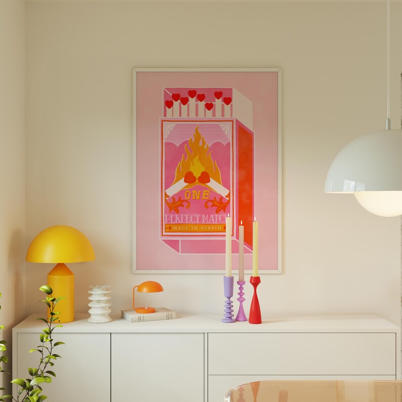 May include: A pink and orange poster with a matchbox illustration. The matchbox has a red flame and the text "ONE PERFECT MATCH MADE IN HEAVEN". Three candlesticks with candles are in front of the poster.