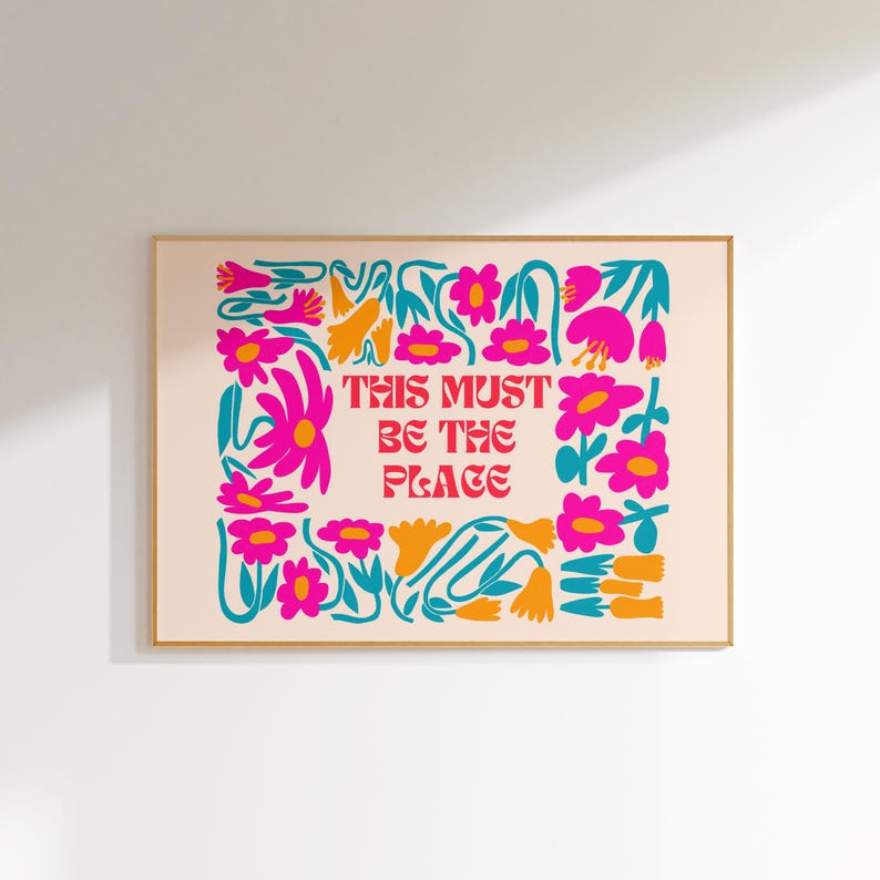 以下が含まれることがあります： ピンクとターコイズブルーの花柄プリントに、赤い文字で「This Must Be The Place」と書かれています。