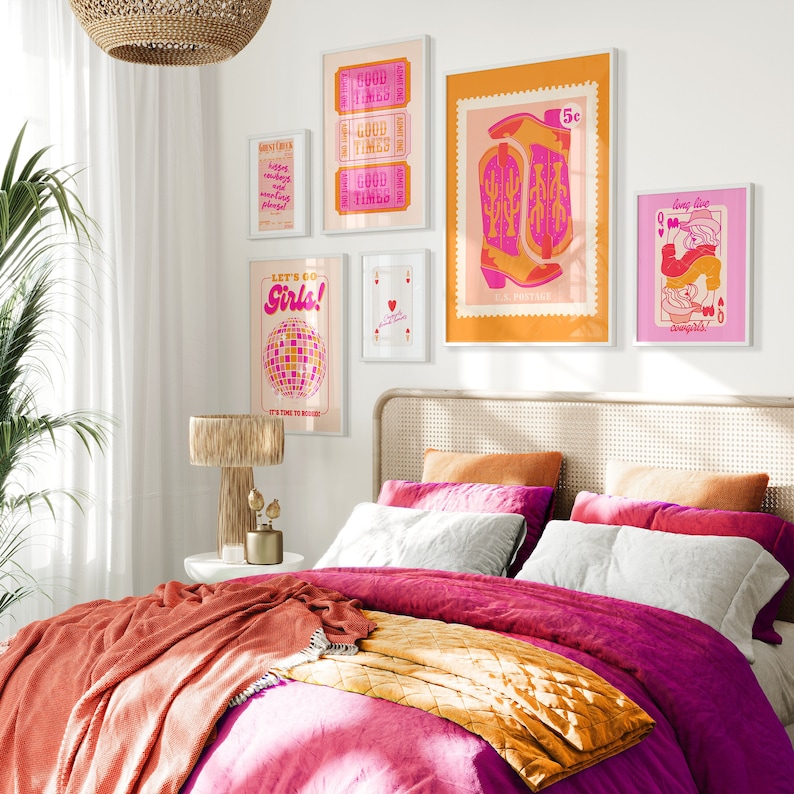 May include: A bedroom with a pink and orange color scheme. The bed has a pink duvet and a yellow throw blanket. There are several framed prints on the wall, including one with the text "Let's Go Girls!" and another with a pair of pink cowboy boots.