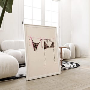 May include: A framed print of a leopard print bikini hanging on a clothesline with pink string. The background is a light beige color.