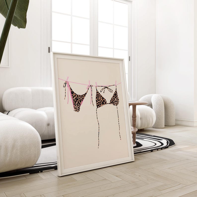 May include: A framed print of a leopard print bikini hanging on a clothesline with pink string. The background is a light beige color.