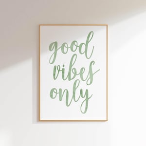 Op de afbeelding: Een ingelijste print met de tekst "good vibes only" in een zachtgroen script. De tekst staat centraal op een witte achtergrond en de lijst is lichtbruin. Het kunstwerk wordt op een witte muur getoond.