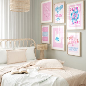 Puede incluir: Un dormitorio con una colcha rosa claro y una cabecera de mimbre. La pared es verde claro y tiene varias impresiones enmarcadas con ilustraciones rosas y azules. Las impresiones incluyen una copa de c&oacute;ctel, una carta de juego y una bolsa de la compra con el texto "&iexcl;Gracias por venir!" Las impresiones tambi&eacute;n incluyen el texto "Good Times" y "Good for the Plot".