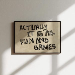 Pode incluir: Uma impress&atilde;o art&iacute;stica emoldurada com o texto "ACTUALLY IT IS ALL FUN AND GAMES" em letras pretas, estilo manuscrito. O fundo &eacute; bege e a moldura &eacute; preta. A obra de arte &eacute; exibida em uma parede branca.