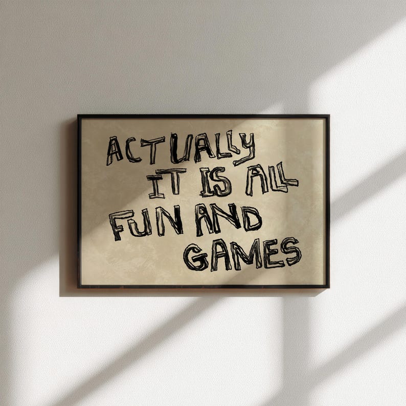 Pode incluir: Uma impress&atilde;o art&iacute;stica emoldurada com o texto "ACTUALLY IT IS ALL FUN AND GAMES" em letras pretas, estilo manuscrito. O fundo &eacute; bege e a moldura &eacute; preta. A obra de arte &eacute; exibida em uma parede branca.