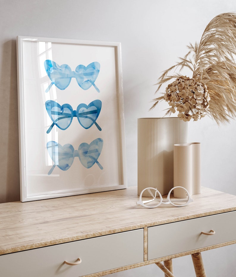 May include: A white framed print with a watercolor illustration of three pairs of blue heart-shaped sunglasses. The print is on a wooden desk with two drawers and a pair of white round glasses on the desk.