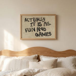 Pode incluir: Uma impress&atilde;o emoldurada com o texto "ACTUALLY IT IS ALL FUN AND GAMES" em letras pretas, estilo manuscrito. A impress&atilde;o tem um fundo bege e uma moldura de madeira marrom clara. A imagem est&aacute; acima de uma cama com travesseiros brancos e uma cabeceira de madeira.