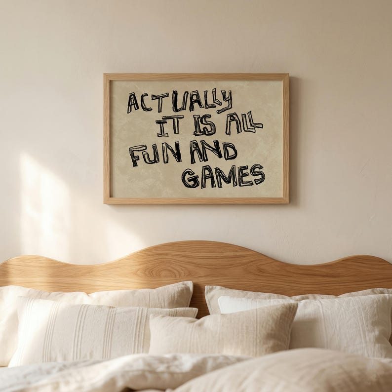 Pode incluir: Uma impress&atilde;o emoldurada com o texto "ACTUALLY IT IS ALL FUN AND GAMES" em letras pretas, estilo manuscrito. A impress&atilde;o tem um fundo bege e uma moldura de madeira marrom clara. A imagem est&aacute; acima de uma cama com travesseiros brancos e uma cabeceira de madeira.