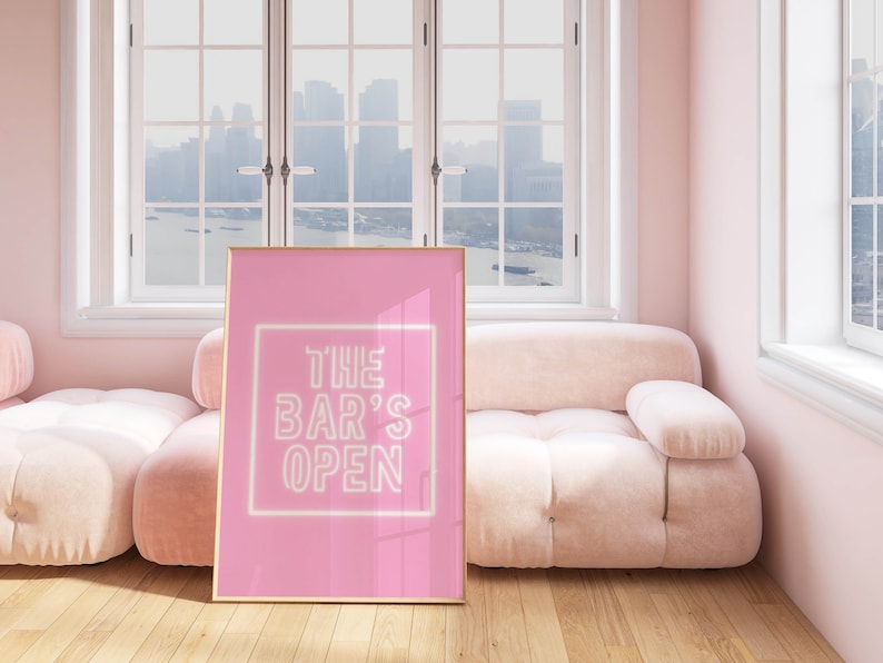 Puede incluir: Un p&oacute;ster enmarcado en rosa con un letrero de ne&oacute;n blanco que dice "THE BAR'S OPEN" en una habitaci&oacute;n rosa con un sof&aacute; rosa y una ventana con vista a la ciudad.