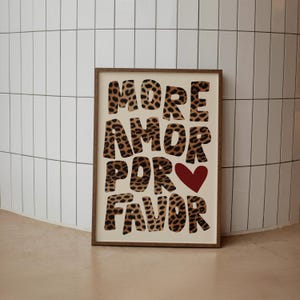 May include: A framed print with the words "More Amor Por Favor" in a leopard print font. A red heart is in the middle of the print.