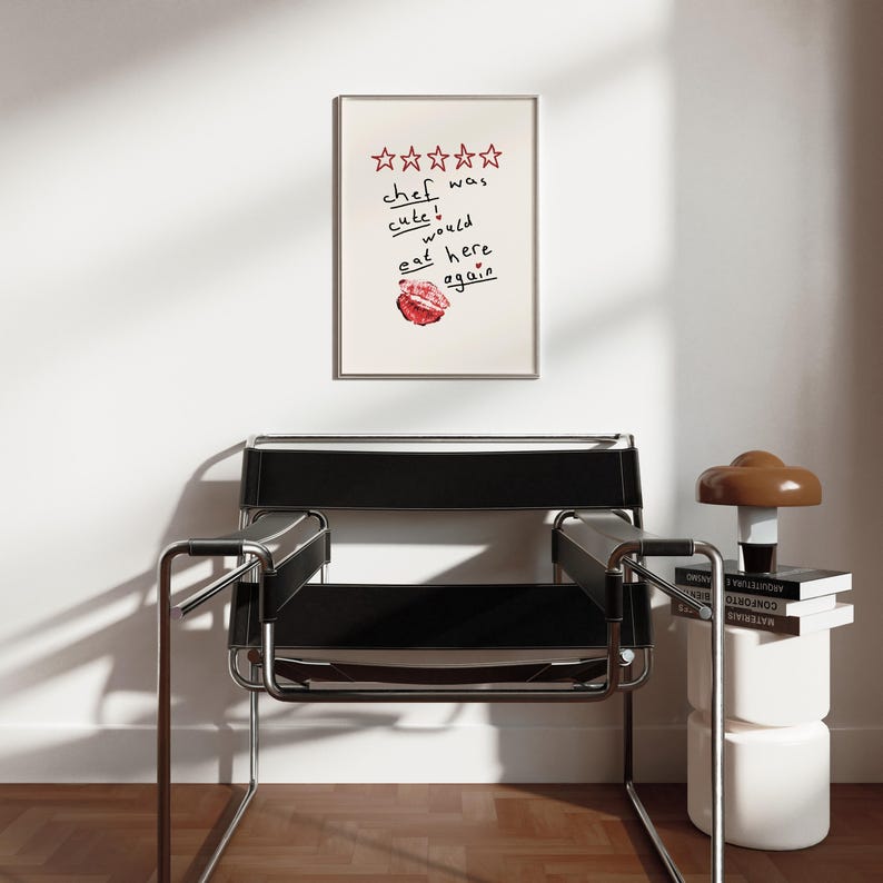 Op de afbeelding: Een ingelijst print met een vijfsterrenbeoordeling en de tekst "kok was schattig, zou hier weer eten". De print hangt aan een witte muur boven een zwarte leren stoel.