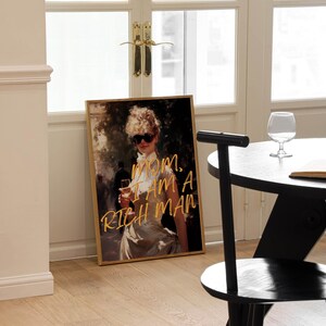 May include: A framed print of a woman in a white dress with sunglasses and a glass of wine. The text "MOM, I AM A RICH MAN" is written in gold lettering on the print.