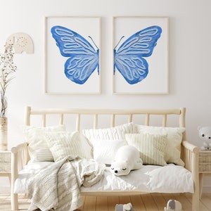 May include: Two framed prints of a blue butterfly with its wings spread wide. The prints are hung on a white wall above a wooden daybed with white bedding and pillows.