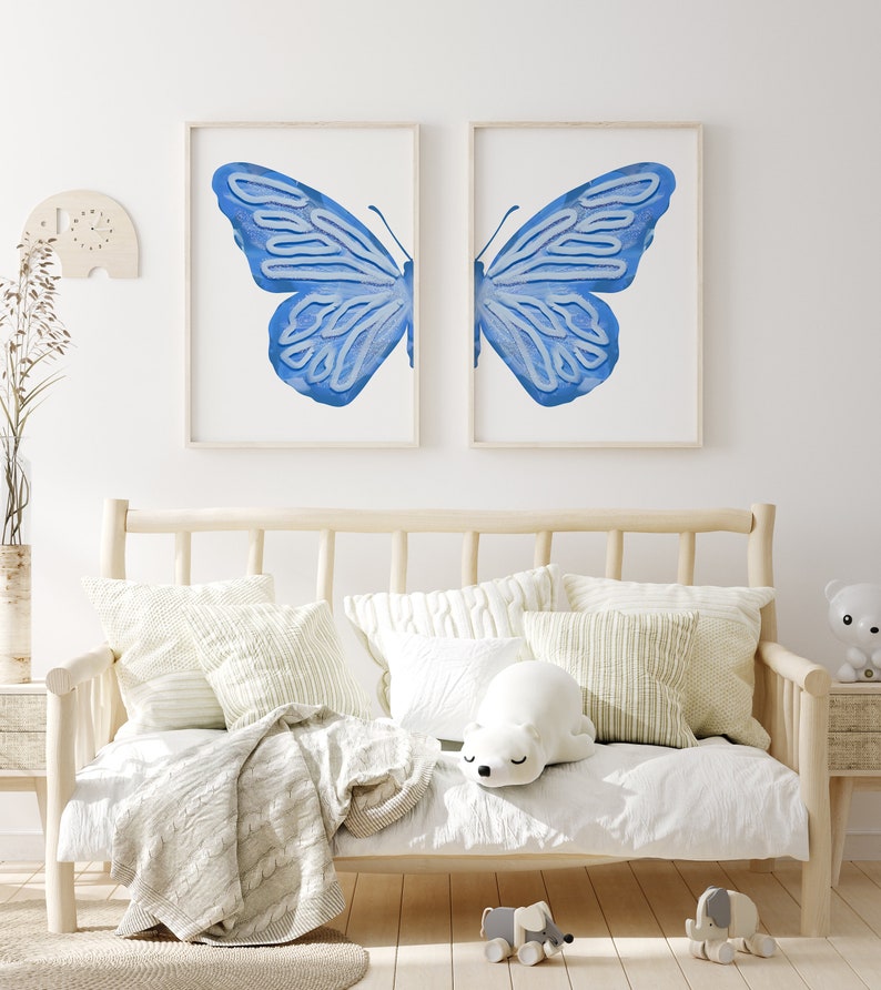 May include: Two framed prints of a blue butterfly with its wings spread wide. The prints are hung on a white wall above a wooden daybed with white bedding and pillows.