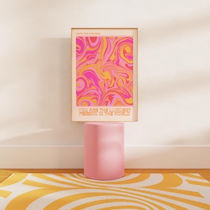Peut inclure: Une impression abstraite rose et orange avec le texte "Trust the Universe" et "You are the luckiest person in the world". L'impression est encadr&eacute;e et expos&eacute;e sur un pi&eacute;destal rose.