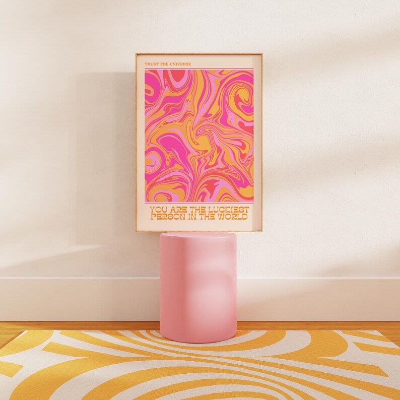 Peut inclure: Une impression abstraite rose et orange avec le texte "Trust the Universe" et "You are the luckiest person in the world". L'impression est encadr&eacute;e et expos&eacute;e sur un pi&eacute;destal rose.