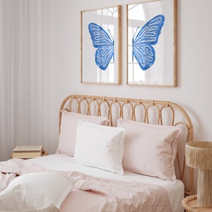 May include: Two framed prints of a blue butterfly with its wings spread wide. The prints are hanging on a white wall above a bed with a light pink linen headboard and bedding.