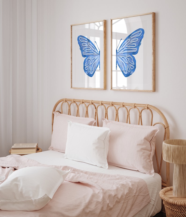 May include: Two framed prints of a blue butterfly with its wings spread wide. The prints are hanging on a white wall above a bed with a light pink linen headboard and bedding.