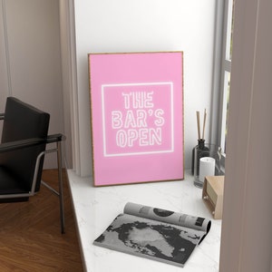 Puede incluir: Una impresi&oacute;n enmarcada rosa con el texto "THE BAR'S OPEN" en letras de ne&oacute;n blancas sobre un fondo rosa.