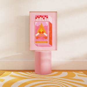 May include: A pink and orange print of a matchbox with the text "One Perfect Match" and a burning match with hearts on the ends. The print is framed and sitting on a pink pedestal.