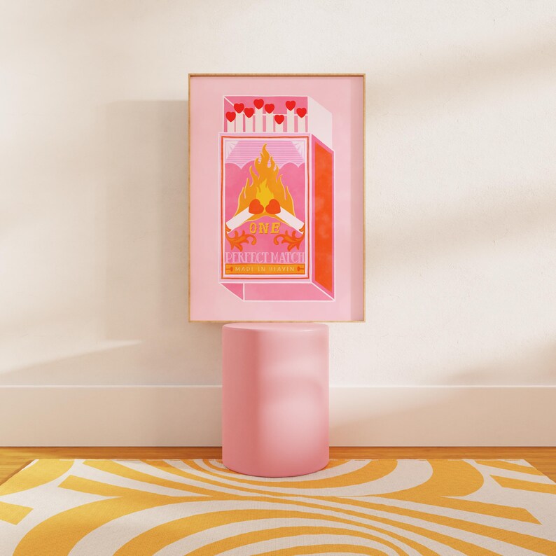 May include: A pink and orange print of a matchbox with the text "One Perfect Match" and a burning match with hearts on the ends. The print is framed and sitting on a pink pedestal.