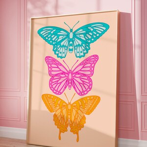 Pode incluir: Uma impress&atilde;o emoldurada com tr&ecirc;s borboletas coloridas em um fundo p&ecirc;ssego. As borboletas est&atilde;o sobrepostas, a borboleta superior &eacute; azul e verde, a borboleta do meio &eacute; rosa e roxa, e a borboleta inferior &eacute; laranja e amarela.