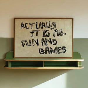 Pode incluir: Uma impress&atilde;o emoldurada com o texto "ACTUALLY IT IS ALL FUN AND GAMES" em preto, exibida acima de uma prateleira verde. A impress&atilde;o tem um fundo bege claro e uma moldura marrom. A prateleira &eacute; verde com borda de madeira natural.