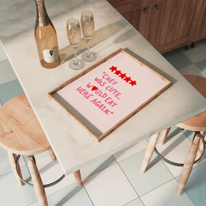 May include: A framed pink poster with the text "Chef was cute, would eat here again." in red lettering with five red stars and a red heart. The poster is on a white countertop with two champagne glasses and a bottle of champagne.