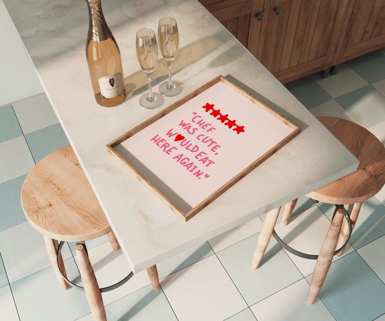 May include: A framed pink poster with the text "Chef was cute, would eat here again." in red lettering with five red stars and a red heart. The poster is on a white countertop with two champagne glasses and a bottle of champagne.