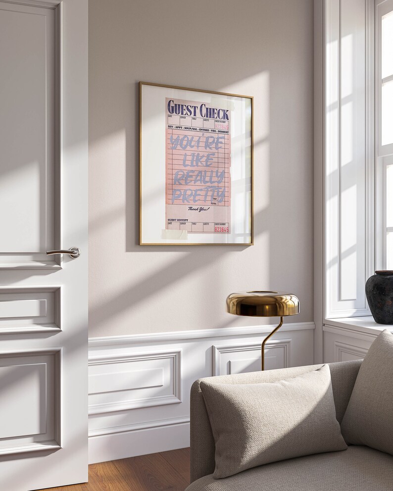 K&ouml;nnte beinhalten: Ein gerahmter Druck eines rosa Gastchecks mit dem Text "You're like really pretty" in blau. Der Druck h&auml;ngt an einer wei&szlig;en Wand in einem Raum mit einem Sofa und einer Lampe.