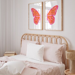May include: Two framed prints of a pink and orange butterfly with its wings spread wide. The prints are hanging on a white wall above a bed with a light pink linen headboard and bedding.