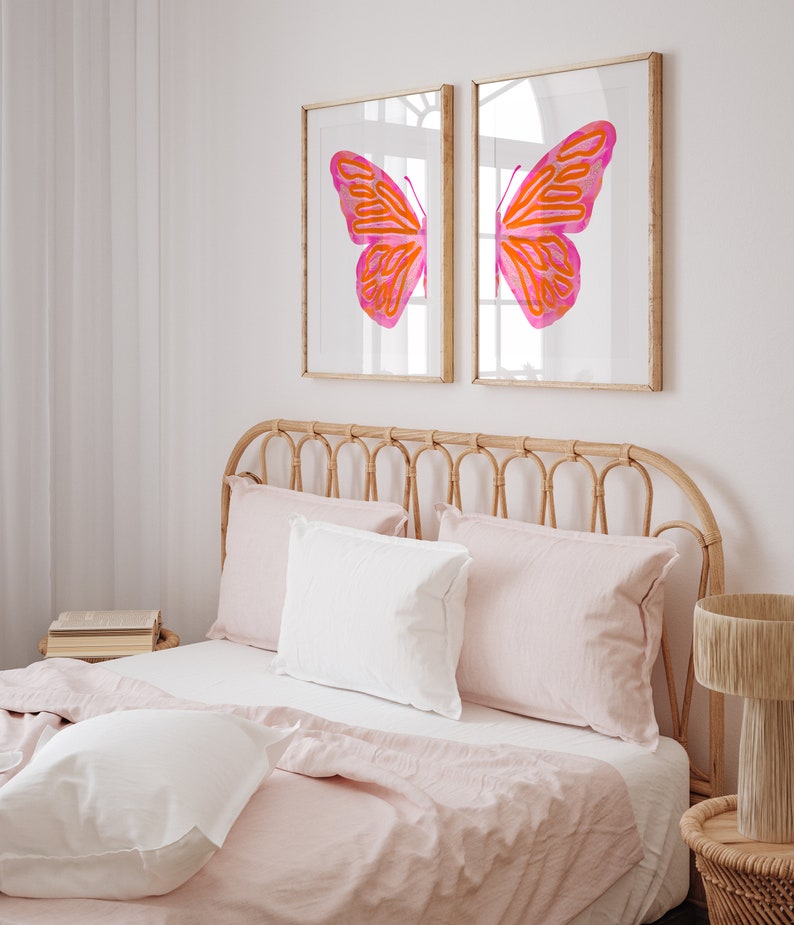 May include: Two framed prints of a pink and orange butterfly with its wings spread wide. The prints are hanging on a white wall above a bed with a light pink linen headboard and bedding.