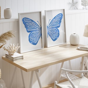 May include: Two white framed prints of blue butterfly wings on a white wall above a light wood desk with a white chair and white decor.