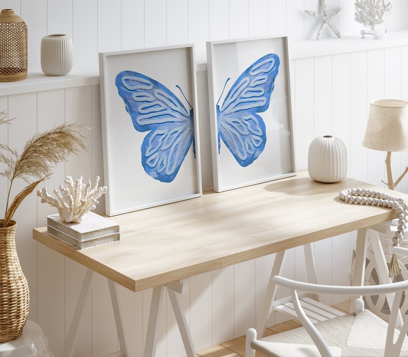 May include: Two white framed prints of blue butterfly wings on a white wall above a light wood desk with a white chair and white decor.