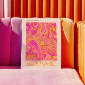 Peut inclure: Une affiche blanche avec un motif abstrait en spirale rose, orange et jaune. Le texte "Trust the Universe" est en haut et "You are the luckiest person in the world" est en bas.