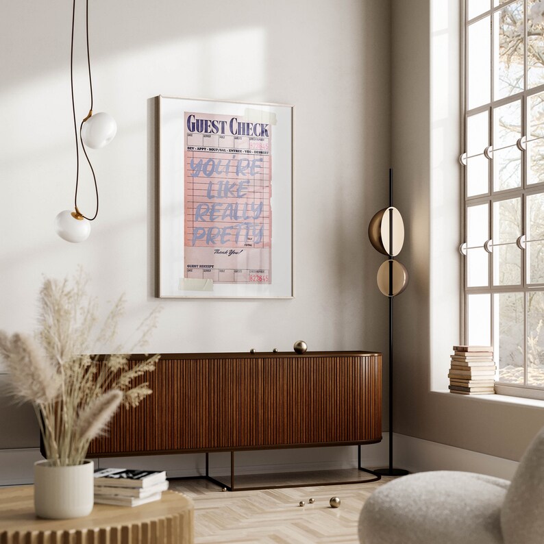 K&ouml;nnte beinhalten: Ein gerahmter Druck eines rosa und wei&szlig;en G&auml;stechecks mit dem Text "You're like really pretty" in lila. Der Druck h&auml;ngt an einer wei&szlig;en Wand &uuml;ber einem Holzschr&auml;nk.