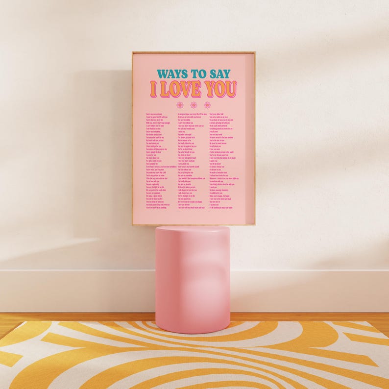 May include: A pink poster with a gold frame that says "Ways to Say I Love You" in a retro font. The poster lists many different ways to say "I love you" in English. The poster is sitting on a pink pedestal.