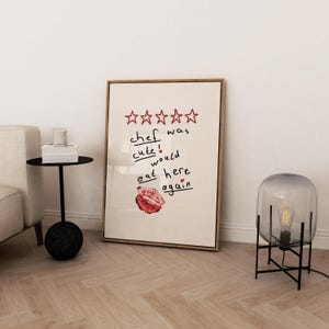 Op de afbeelding: Een ingelijst print met een vijfsterrenbeoordeling en de tekst "kok was schattig! zou hier weer eten". De print hangt aan een witte muur boven een zwarte bijzettafel met een glazen kaarsenhouder en boeken.
